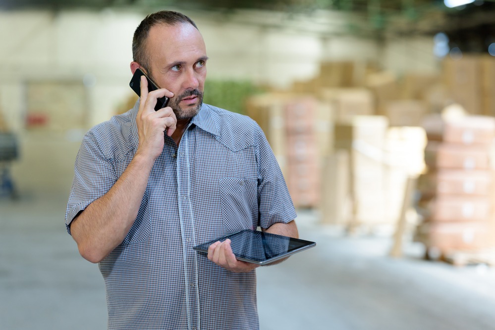 A worker looking at a digital tablet while phoning in warehouse
