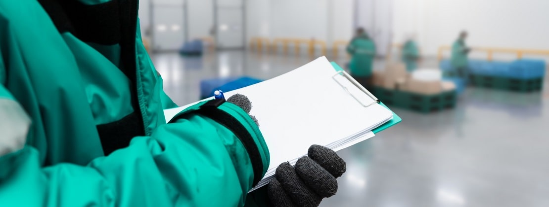 A warehouse worker in a thick jacket holding a clipboard with documents