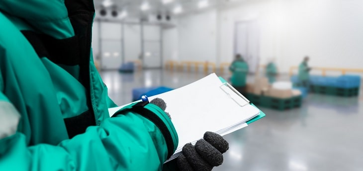 A warehouse worker in a thick jacket holding a clipboard with documents