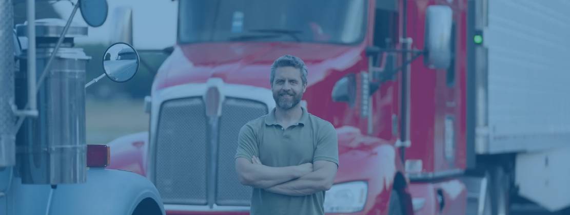 A truck driver with arms crossed smiling in front of a freight truck