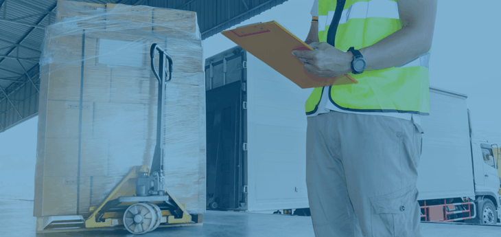 Dock worker looking at clipboard with a stacked pallet on a pallet jack behind him.